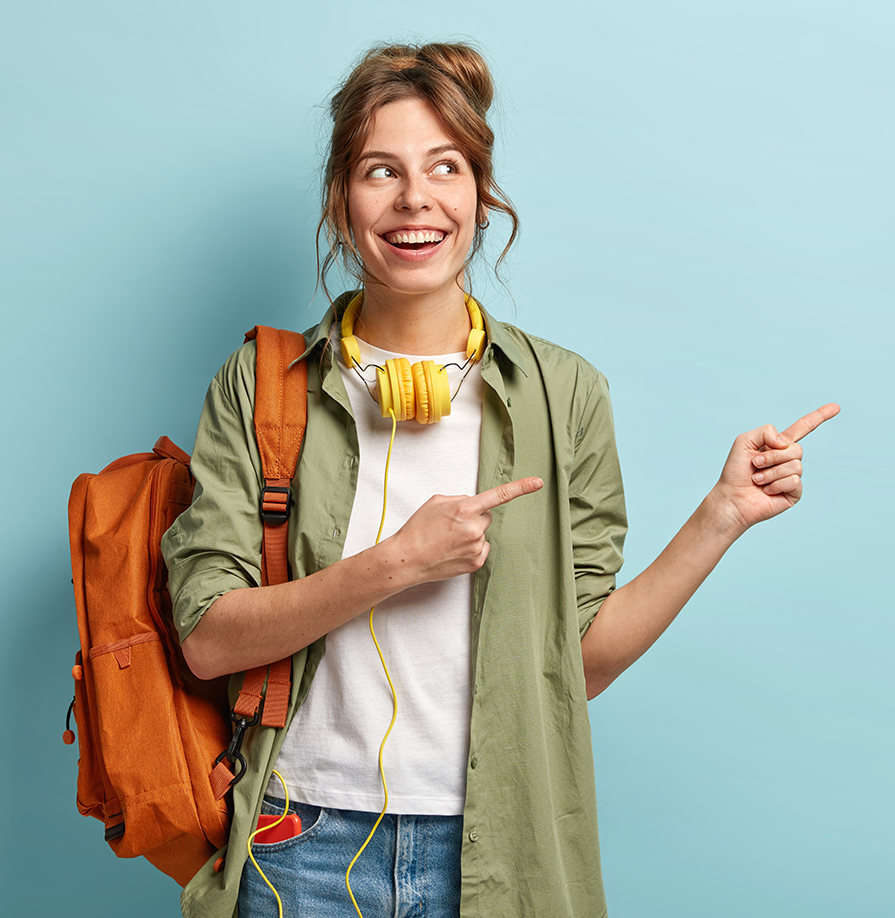 Positive European female teenager uses modern technologies for entertainment, points aside on free space, dressed in loose shirt and jeans, carries backpack, promotos something, poses indoor